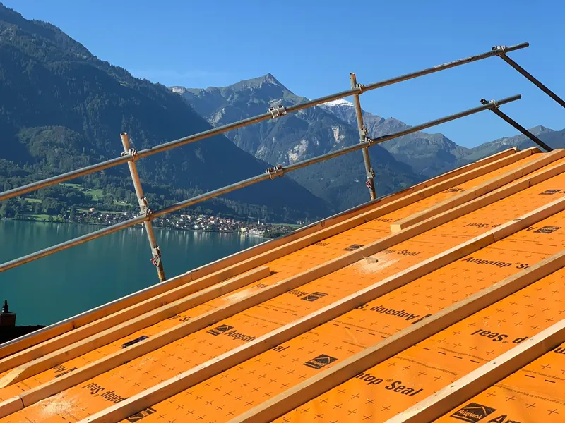 Frisch gelattetes Dach mit Gerüstgeländer, im Hintergrund Berge und See